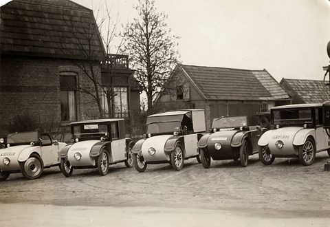 Five small rounded cars with single central headlamps parked in a row before brick houses on a sandy yard.