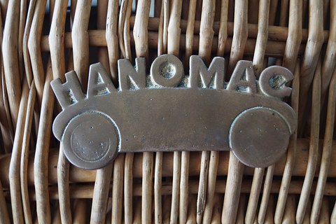 A bronze badge shaped like a small car with the word Hanomag above it, fixed to woven wicker. Opens a larger view