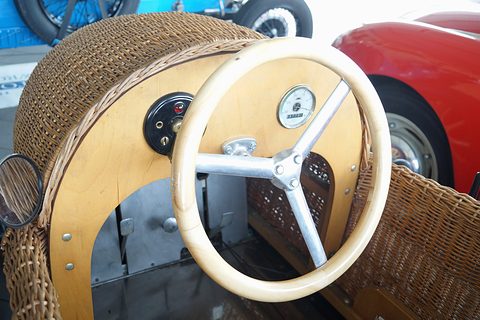 A cream steering wheel and a plywood dash with one gauge, framed by a woven wicker body. Opens a larger view