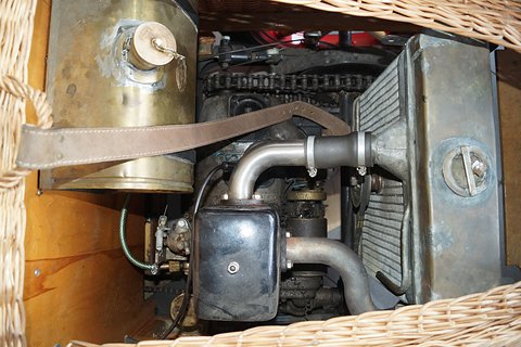 A brass fuel tank on a leather strap above a small engine and a finned radiator inside a wicker body. Opens a larger view