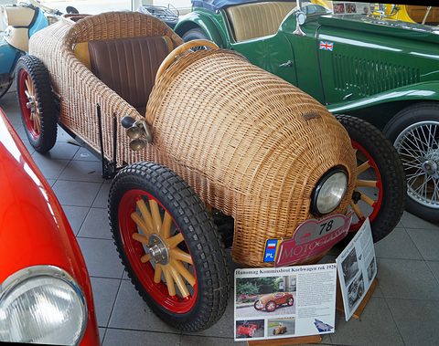 A small car whose rounded body is woven from wicker, with red wooden wheels and a single headlamp, by a museum board. Opens a larger view