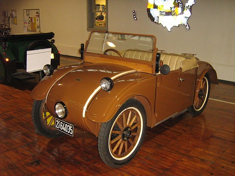 A brown open two-seater with cream coachlines, a single headlamp and wooden wheels on a wooden floor. Opens a larger view