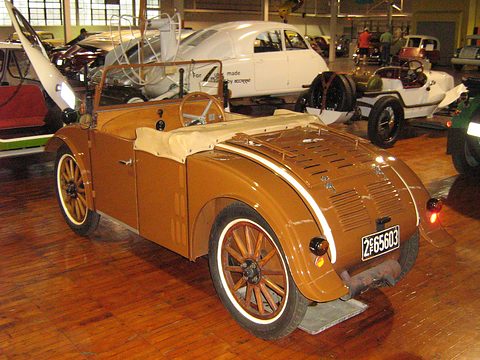 Rear three-quarter view of a brown open two-seater with cream coachlines, a louvred tail and wooden wheels. Opens a larger view