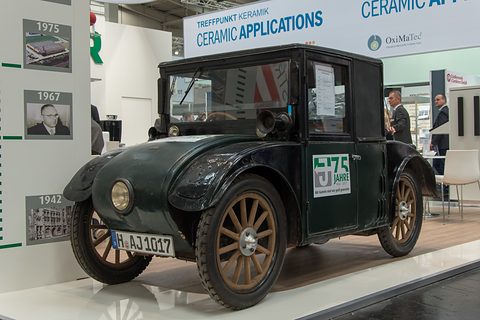 A small dark green saloon with a tall glazed cab, wooden wheels and a single headlamp on a trade-fair stand. Opens a larger view
