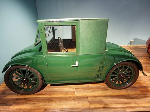 Side view of a small green saloon with a tall cab, a single door and green wooden wheels on a wooden floor. Opens a larger view