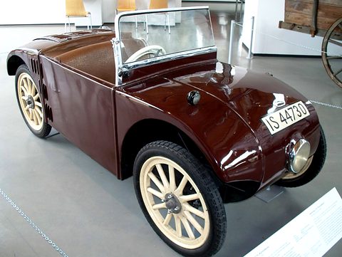 A maroon open two-seater with cream spoked wheels, a single headlamp and plate IS-44730 on a grey floor. Opens a larger view