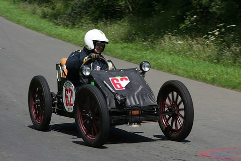 A helmeted driver in a tiny bare-wheeled racing car with a race number on its nose rounds a bend. Opens a larger view
