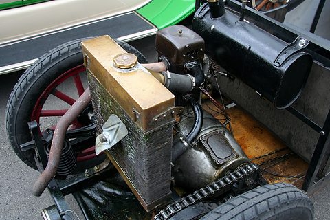 Close view of a brass-topped radiator, a single-cylinder engine and a drive chain on a bare chassis. Opens a larger view