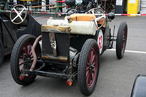 A stripped racing car with a radiator at the front, a long exhaust pipe, wooden wheels and race number 62.