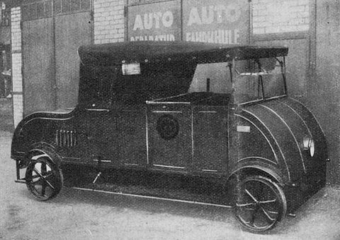 A half-tone of a slab-sided small car with a canvas roof standing on rail wheels before a garage wall. Opens a larger view