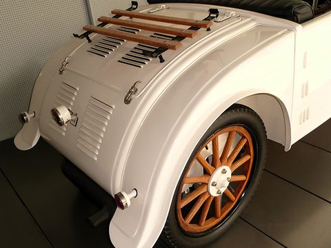 The rounded cream tail of a small car with rows of louvres, a slatted wooden rack and an orange wooden wheel. Opens a larger view