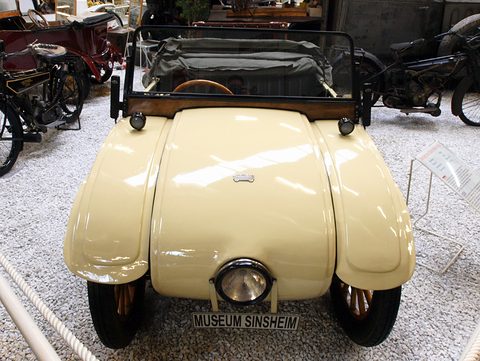 Front view of a cream rounded car with a single headlamp and a Museum Sinsheim plate on a white gravel floor. Opens a larger view