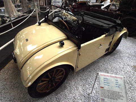 A cream open two-seater with wooden wheels on white gravel, a museum information board in front. Opens a larger view