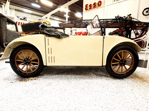 Side view of a cream two-seater with wooden wheels and a folded hood on white gravel, motorcycles behind. Opens a larger view