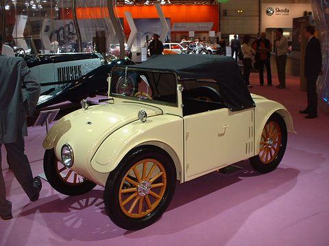 A cream two-seater with a black hood, wooden wheels and a single headlamp on a pink show carpet. Opens a larger view