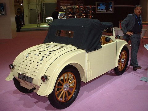 Rear three-quarter view of a cream two-seater with a louvred rounded tail, a black hood and wooden wheels. Opens a larger view
