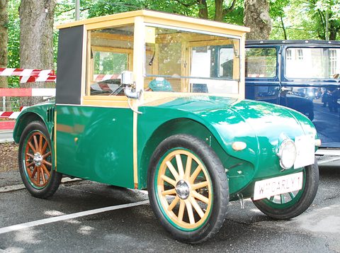 A small green saloon with a light wooden-framed glass cab and wooden wheels parked on a street under trees. Opens a larger view