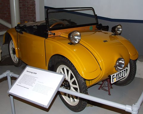 A yellow open two-seater with white spoked wheels and a single central headlamp behind a museum label. Opens a larger view