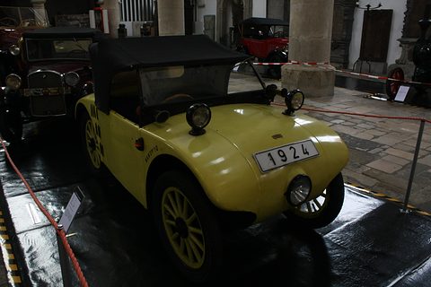A yellow two-seater with a black hood, yellow wheels and a 1924 plate behind a red rope on a dark floor. Opens a larger view
