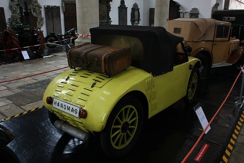 Rear three-quarter view of a yellow two-seater with a leather suitcase on a rack and a Hanomag plate. Opens a larger view