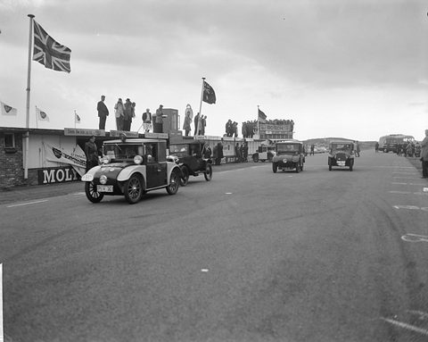 A small dark saloon with a single headlamp drives down a wide track ahead of other old cars, flags overhead. Opens a larger view