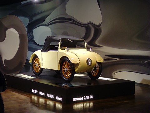 A cream two-seater with a brown hood and orange wooden wheels on a black plinth before a swirling grey mural. Opens a larger view