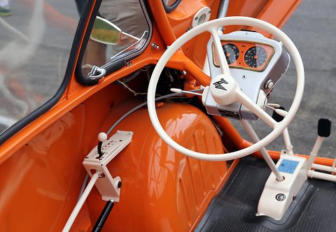 Cream two-spoke steering wheel in front of orange and black dials, a white gear gate on the floor beside it. Opens a larger view