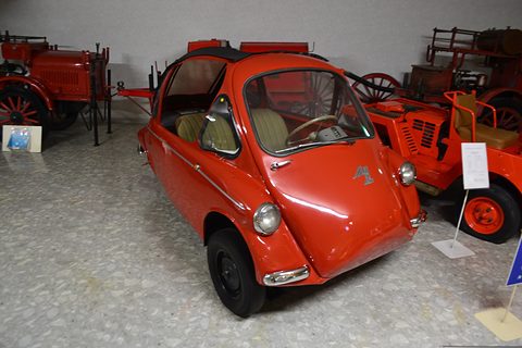 Orange bubble car on a terrazzo floor among red vintage fire engines. Opens a larger view