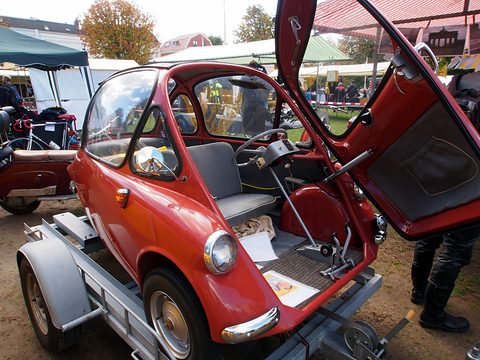 Red bubble car on a trailer with its front door open, showing grey bench seat, steering column and pedals. Opens a larger view