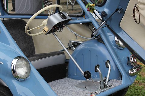 Pale blue cabin with a cream wheel on a column, dark seat, floor pedals and a chrome headlamp in the foreground. Opens a larger view
