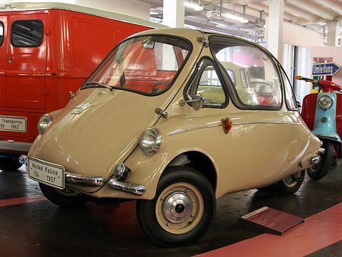Cream bubble car with a ‘Heinkel Kabine 154 1957’ display plate, a red van and a scooter behind. Opens a larger view