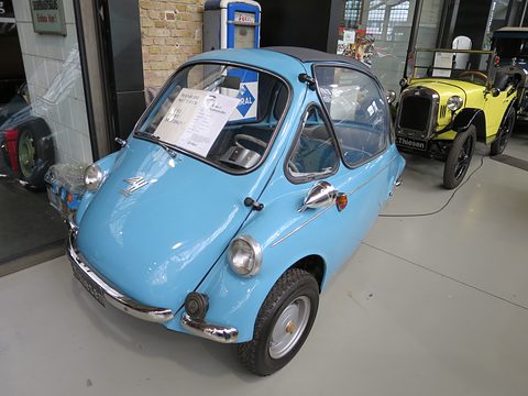 Sky-blue bubble car parked indoors on a pale floor, a yellow vintage roadster and a fuel pump behind. Opens a larger view