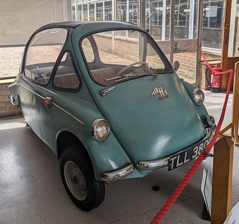 Faded green bubble car behind a red rope in a glazed museum hall, registration TLL 380. Opens a larger view