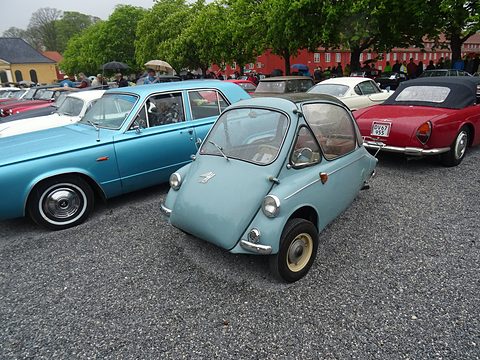 Pale blue bubble car on gravel between a turquoise American saloon and a red roadster. Opens a larger view