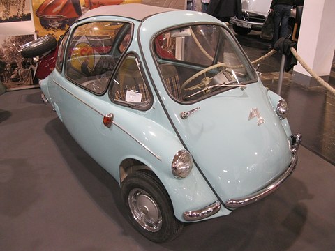 Pale blue bubble car with a white roof on a dark show floor, period photographs on the wall behind. Opens a larger view