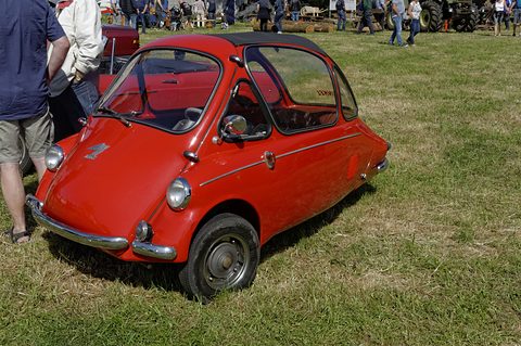 Red bubble car parked on dry grass, visitors and a tractor in the field behind. Opens a larger view