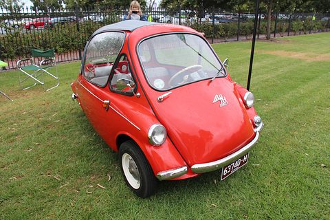 Red bubble car on a mown lawn, folding chairs and a fence of parked cars behind. Opens a larger view