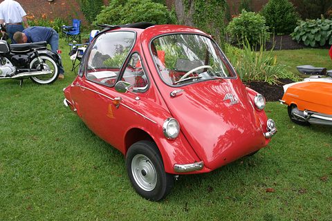Red bubble car on grass in a garden, a motorcycle and an orange microcar alongside. Opens a larger view