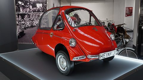 Red bubble car with a HEINKEL name plate on a lit platform, a large period crowd photograph behind. Opens a larger view