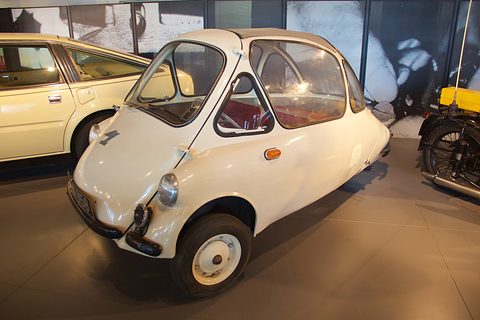 Cream bubble car on a polished floor beside a pale estate car and a motorcycle. Opens a larger view