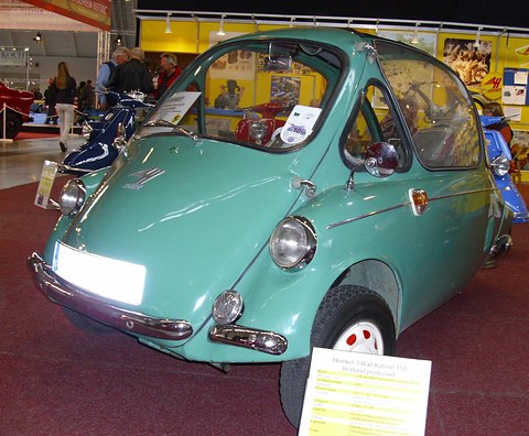 Turquoise bubble car on a red carpet at an indoor show, an information card propped against its front wheel. Opens a larger view