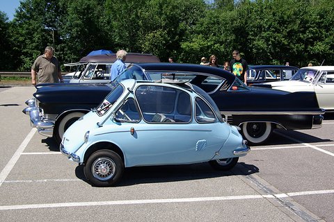 Pale blue bubble car in side profile in front of a black American saloon. Opens a larger view
