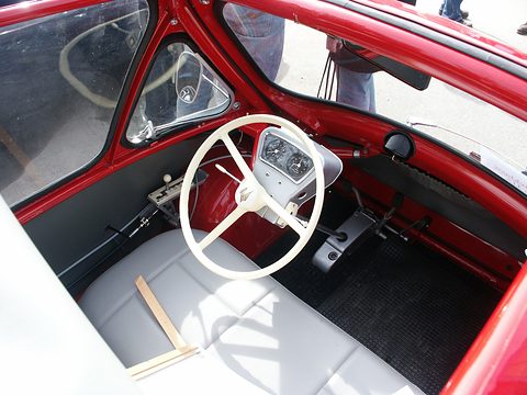 Looking down into a red bubble car: white two-spoke steering wheel, small instrument pod and pale grey bench seat.