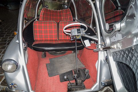 Open front door revealing tartan bench seats, red carpet, a white steering wheel on its column and pedals. Opens a larger view