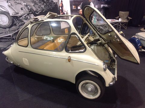 Cream bubble car with its front door raised, showing tan seats, on a dark show carpet. Opens a larger view