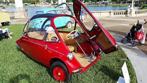 Red bubble car on grass beside a lakeside promenade, its front door swung up, a pram and visitors nearby. Opens a larger view