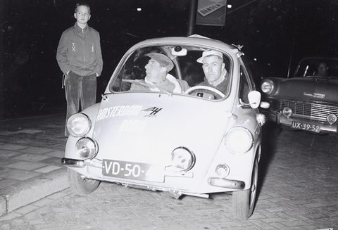 A small pale bubble car seen from the front at night, two men in caps behind its screen, a boy standing at the kerb.