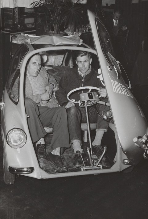 Two men in sweaters on the bench of a bubble car, its front door with the steering wheel swung open, a plant behind.