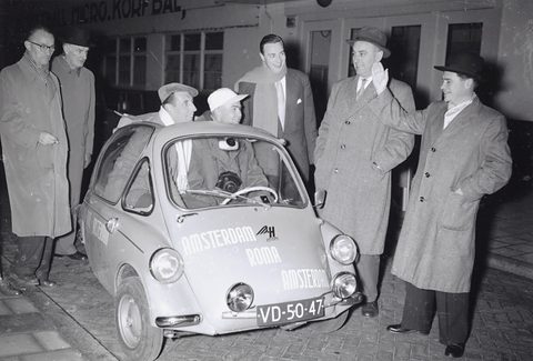 Men in overcoats and hats round a small bubble car with lettering on its nose, one man waving, two men inside.
