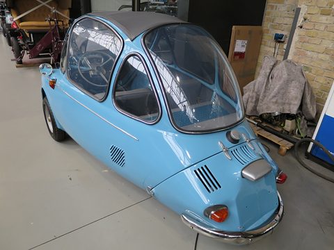 Back of a sky-blue bubble car: engine louvres, small tail lamps and curved chrome bumper, in a brick hall. Opens a larger view